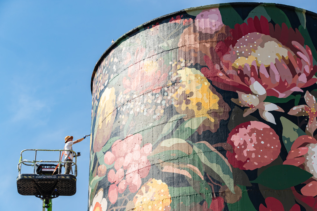An artist on a cherry picker paints a large, colourful floral mural on a tall cylindrical structure under a clear blue sky, showcasing vibrant flowers and lush green foliage, located at Federal Mills North Geelong, Owned by Hamilton Group property specialists