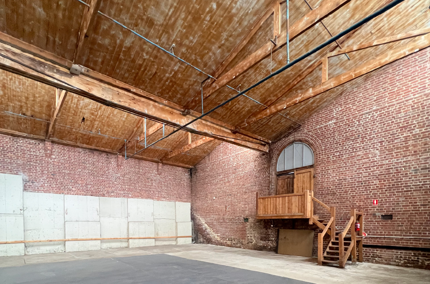 Interior of a historic industrial warehouse with exposed red brick walls, timber trusses, and pitched wooden ceiling, featuring a rustic elevated wooden platform and arched window door. Pegasus Building Geelong