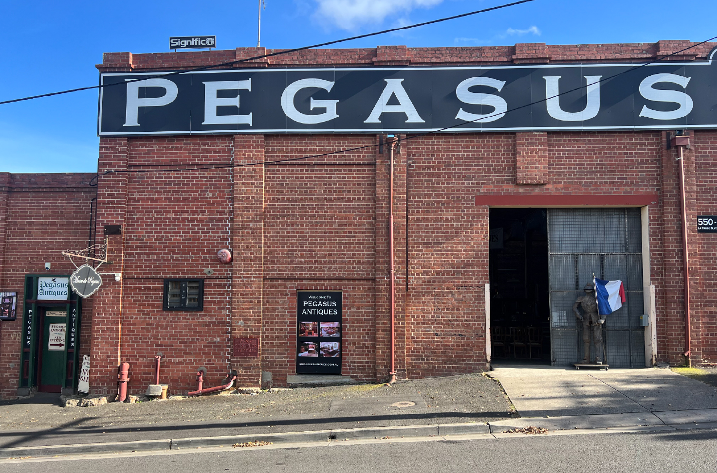 Red brick facade with steel sliding door, old set of armour at the front holding french flag, large black sign with white writing saying "PEGASUS" old concrete path runs across the front of the building, Pegasus Building being renovated by Hamilton Group Geelong