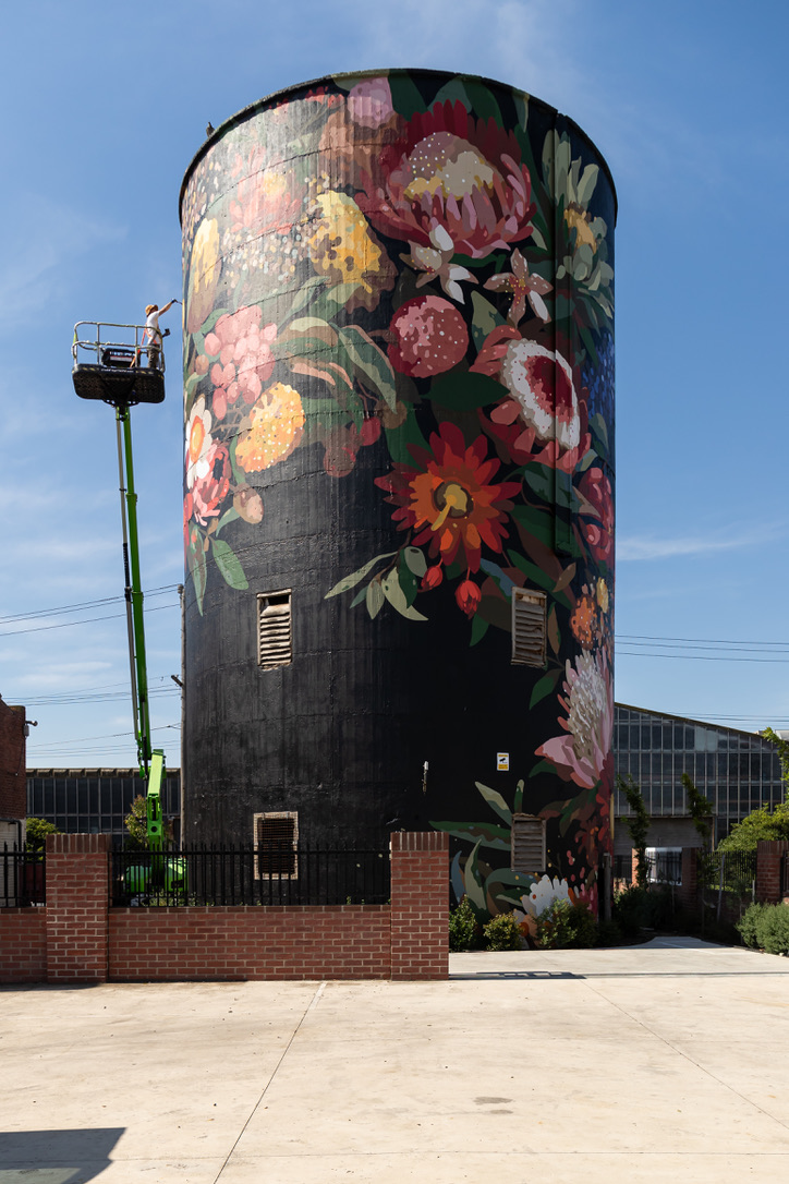 Artist Sai Neoh paints a large floral mural on a cylindrical silo at Federal Mills in North Geelong. The artwork features vibrant, oversized native flowers on a black background. The silo is enclosed by a low brick fence, and adjacent industrial buildings are visible in the background under a clear blue sky.