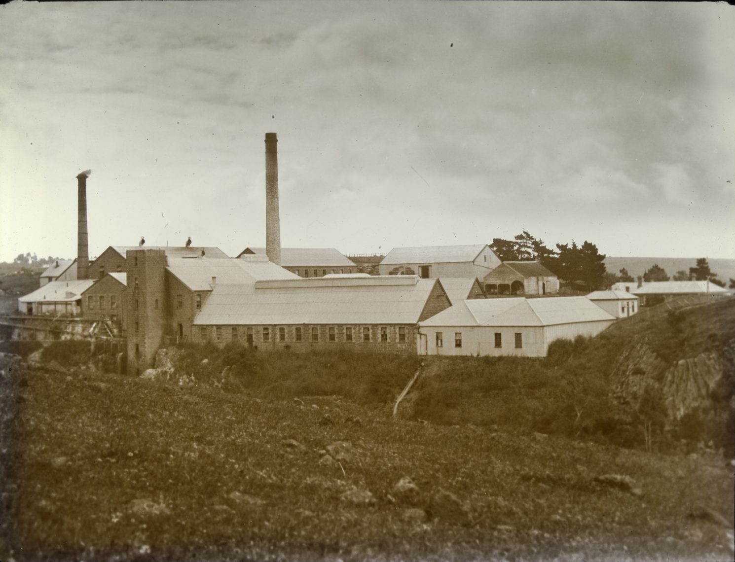 An old photo of the original Fyansford paper mills located in Geelong, The factory is made of multiple smaller sheds, most of the factory is made up of blue stone and red brick walls, two large chimneys stand tall within the precinct, grass land and trees surround the area, Fyansford Paper Mills is being renovated by Hamilton Property Specialists Geelong