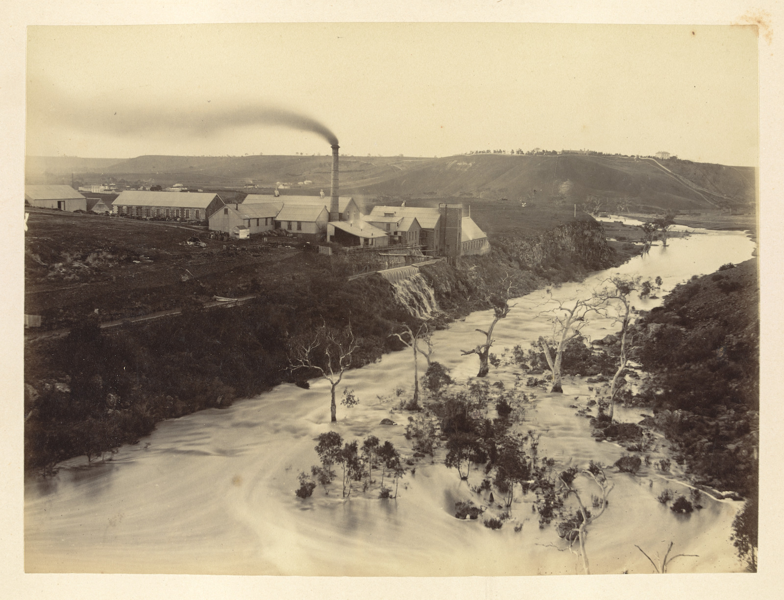 Vintage photo of the Fyansford paper mills during a flood, the large factory is situated on top of a hill with an over flowing river gushing past it, the factory is in full operation with smoke billowing out of a large chimney in the centre, Hamilton Group Geelong