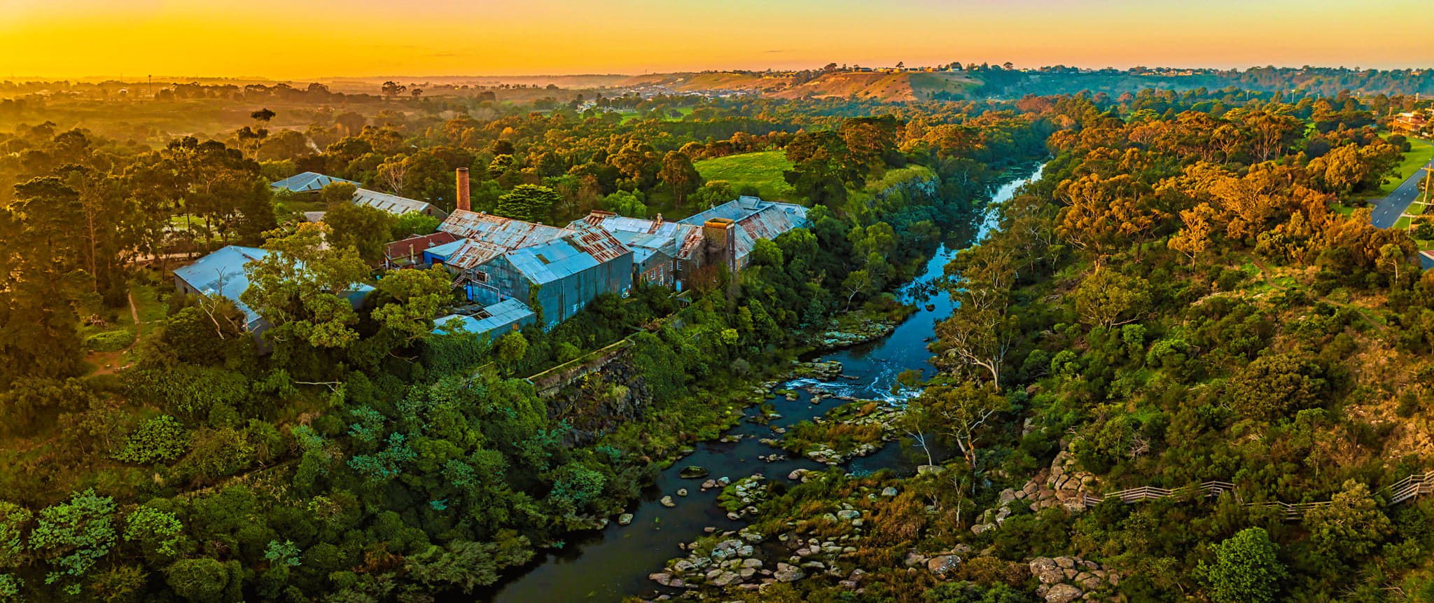 Aerial view of the Fyansford Paper Mills during a sunset, the historic building is surrounded by dense bushland with large trees scattered throughout, a long river winds through the greenery, off in the distance is some hills wi houses atop them, Fyansford Paper Mills is now owned by the Hamilton Property Group Geelong