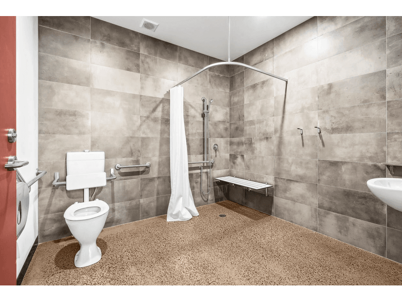 Ambulant bathroom space with large brown and grey tiles filling the walls, a disabled toilet to the left, with support rails on the wall, to the back right is a shower space with a curtain and a small white bench, and a basin mounted on the wall to the right, Hamilton Group Geelong