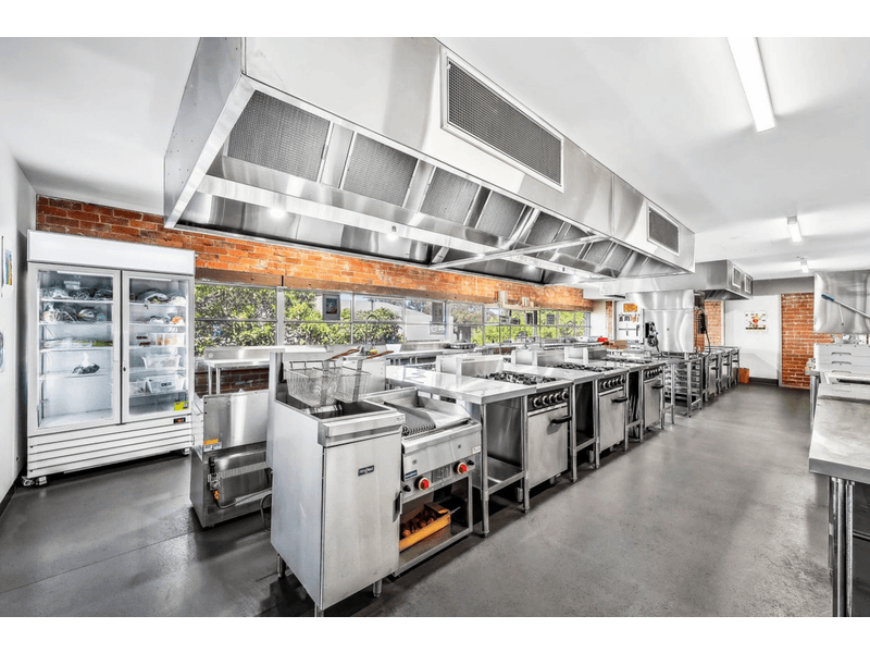 Large industrial kitchen set-up, it features multiple stove tops and ovens and a deep fryer, all in stainless steel, above are large extraction fans, to the left side of the room and a white fridge, next to the is a long set of windows letting in natural light, Hamilton Geelong