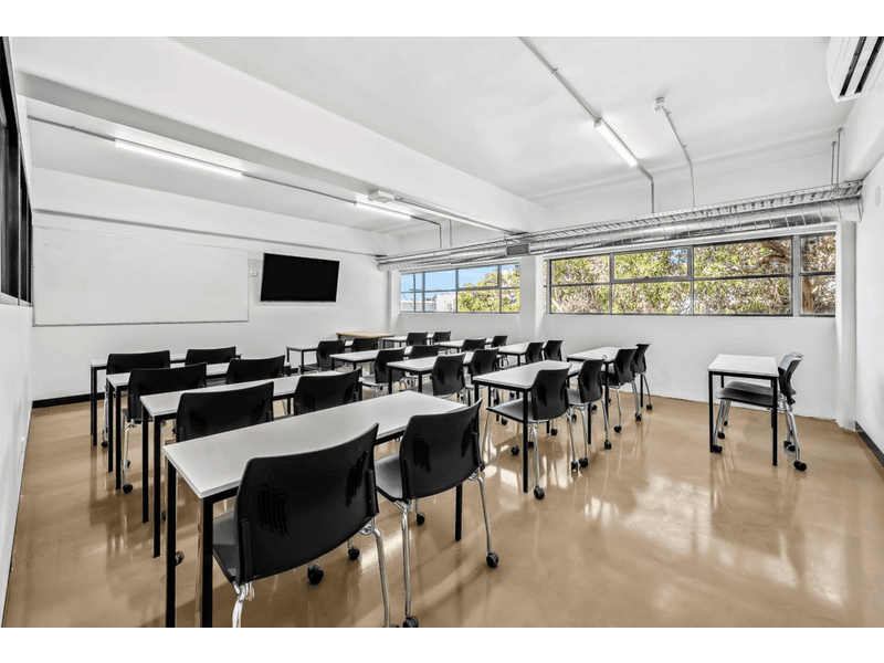 Class room setup featuring many black chairs surrounding small white desks, tan coloured flooring , white walls and roof, large whiteboard on the far wall with a TV next to it, the back wall is filled with windows letting in natural light, Hamilton Property Specialists