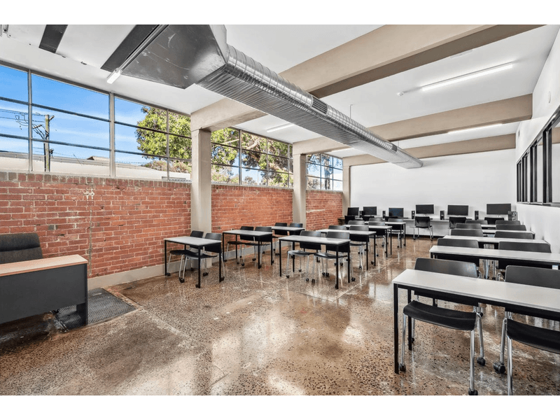 Long classroom featuring many office desks with black chairs, red brick wall to the left side with windows, white walls surround the rest, overhead is a large air con vest, in the front left corner of the room is a desk with an ergonomic chair behind it, Hamilton Group