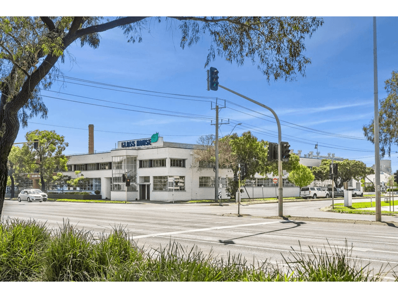 Street view of large intersection with sets of lights, lush greenery surround the roadway, across the road is a large white building with "GLASS HOUSE" printed on the roof, in the distance is a large chimney, above are clear blue skies, Hamilton Group Geelong