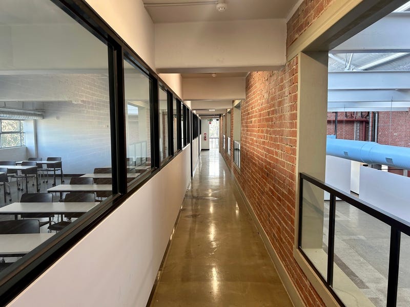 Long red brick hallway with views out into a large warehouse space, each viewing space has a tbar balustrade filled with glass, on the left side is windows that look into a classroom area with many tables and chairs, Hamilton Group Leasing