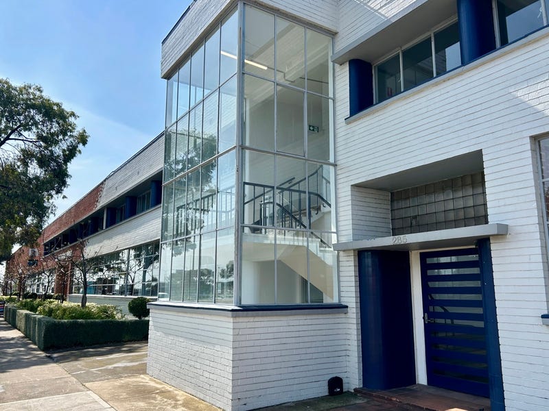 Front view of The Glass House in North Geelong, A white brick facade starts from the right and in the distance it changes to red bricks, in the centre of the shot is a stairwell connecting the two floors, a large bluse door is off to the right with some fancy square glass above it, off to the far left is a well maintained lush green garden, Hamilton Property Group Geelong