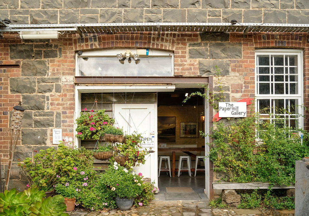 Blue stone and red brick wall filled with an old style white window it has a sign out the front saying "The Papermill Gallery" the building is surrounded by a lush green garden that has many flowers sprouting out, Building owned by Hamilton Property Specialists