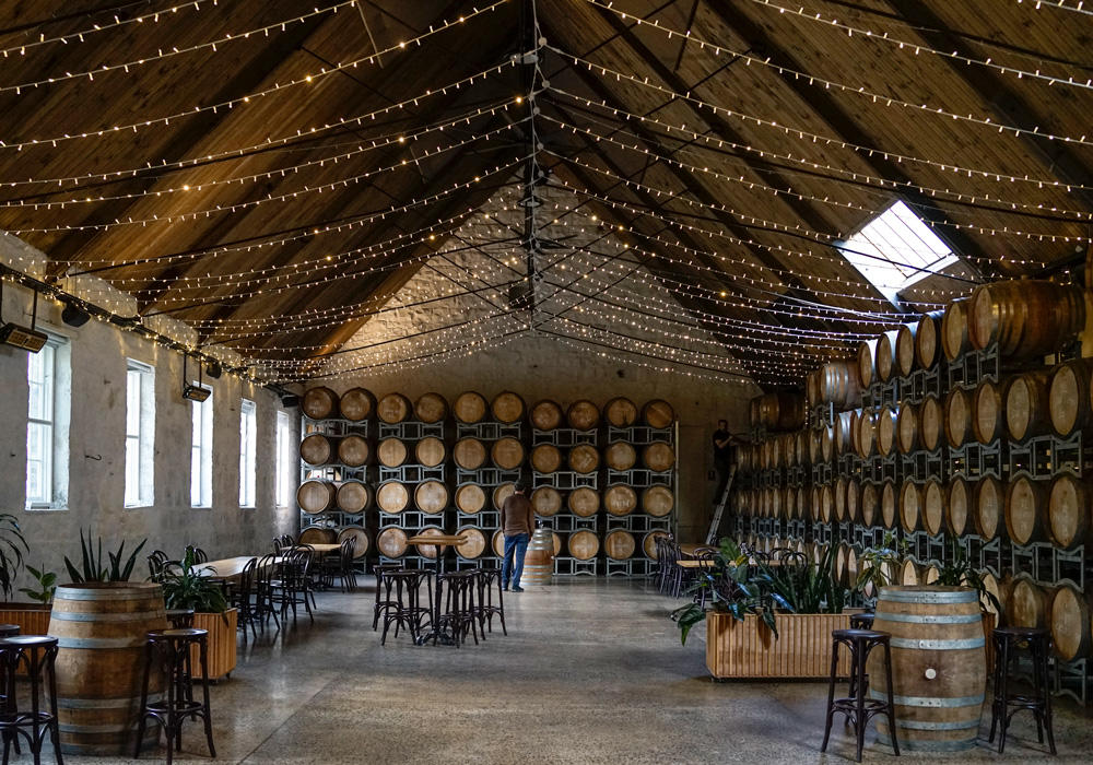 Grand hall surrounded by timber barrels stacked to the roof, a peaked timber roof is held up by thin steel trusses, weaved between the trusses is long sets of fairy lights, chairs and tables sit upon polished concrete floors, Building owned by Hamilton Property Group